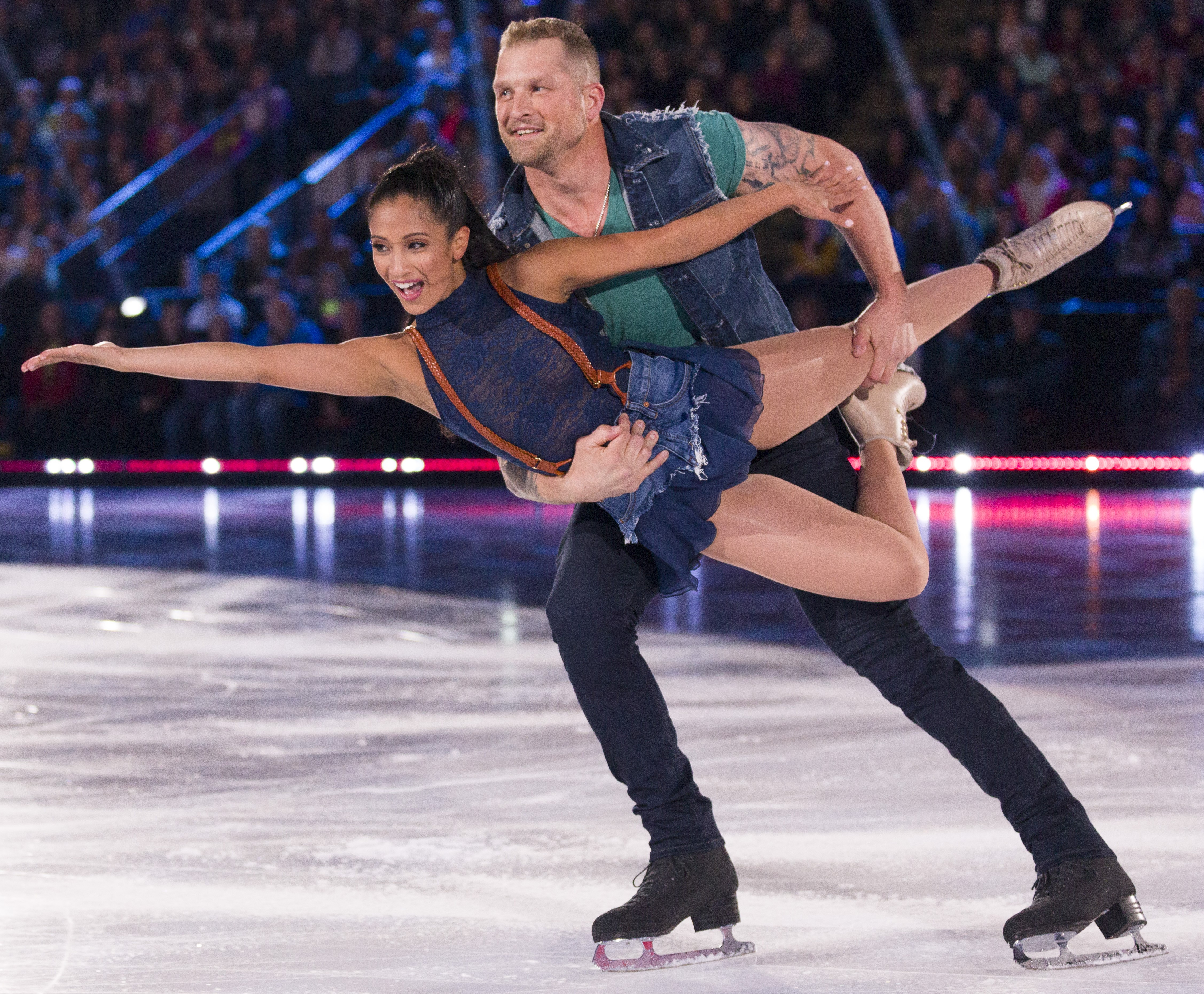 Amanda and hocket player Colton Orr perform on Battle of the Blades. Amanda is wearing a blue top with orange suspenders and a denim skirt. Colton holds Amands up in the air as she holds her arms extended. He is wearing a green shirt with a denim vest and black pants. 