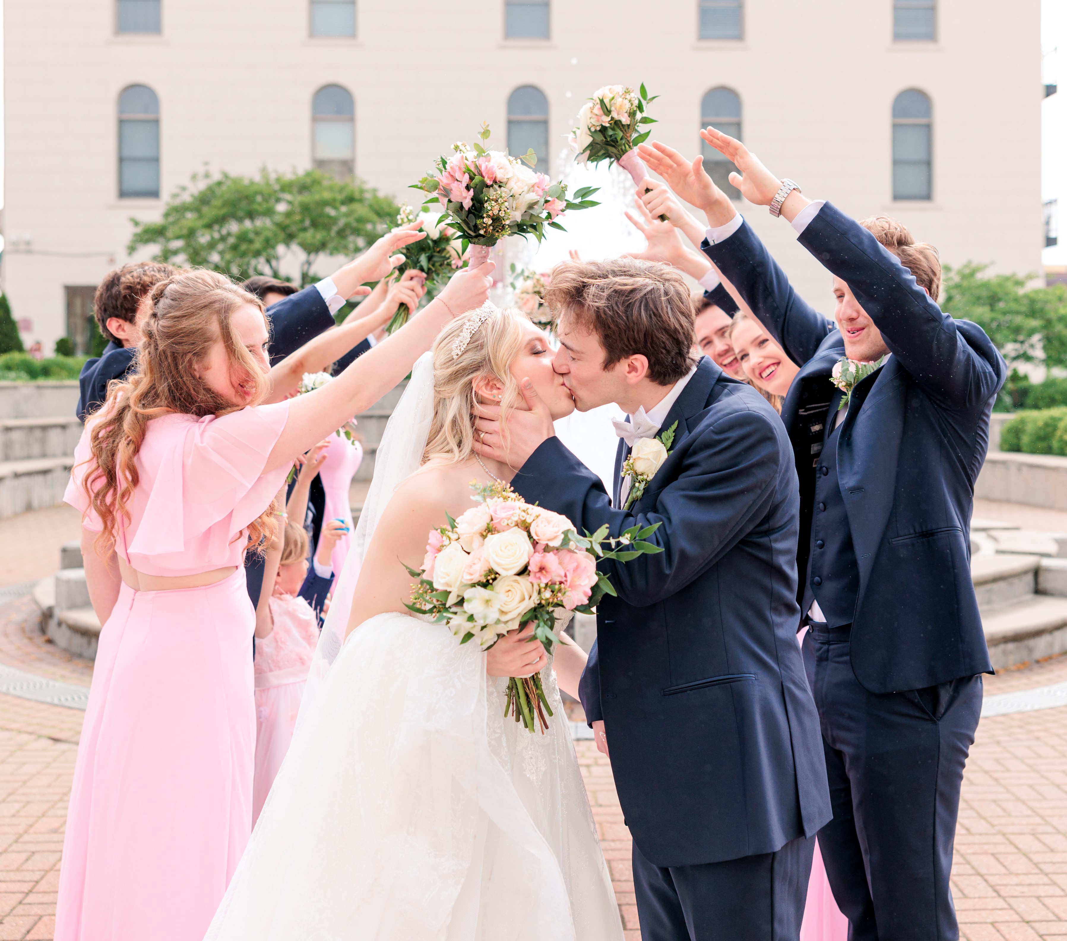 Eva Pate and Logan Bye share a kiss in front of their wedding party, who had formed a tunnell. She wears a white wedding dress, him a navy ux. She holds her bouquet of white flowers.