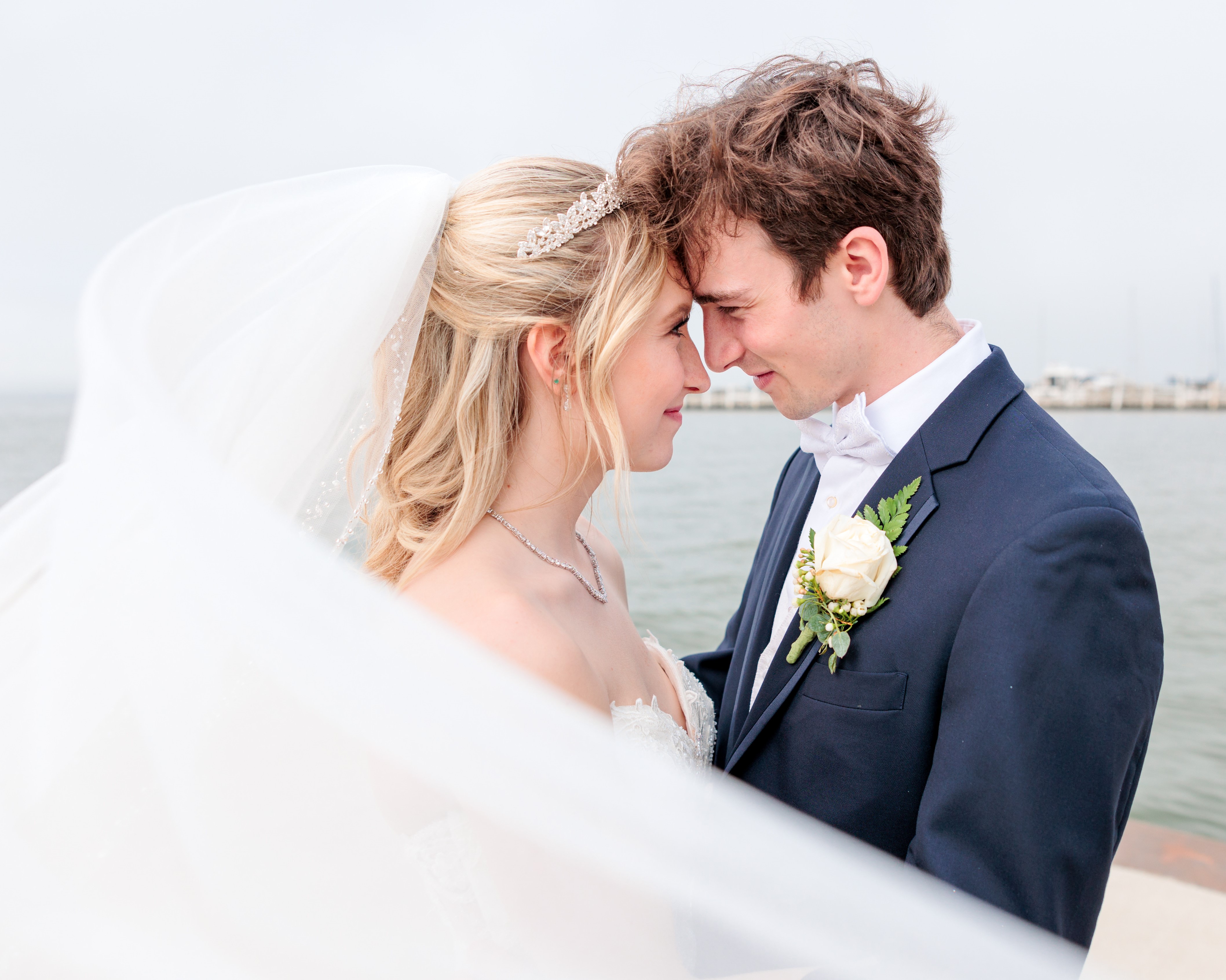 Eva and Logan face each other, their foreheads touching. She wears her wedding dress, him a navy tux. Her veil is blowing in the wind toward the camera, framing the photo.