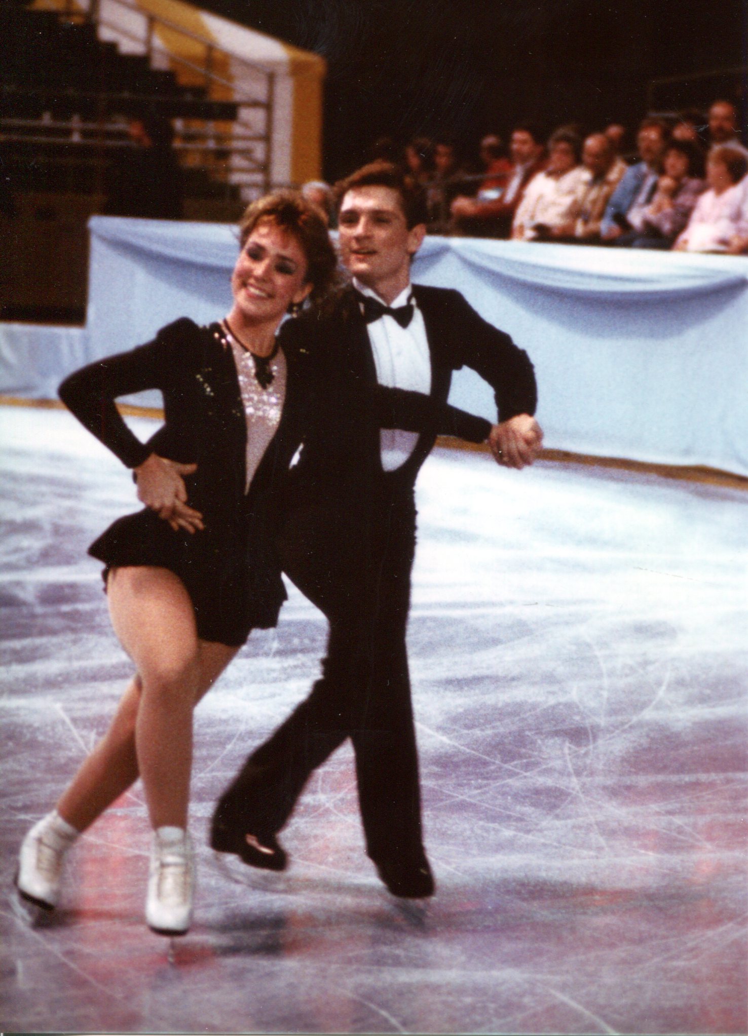 Susan and Joe compete wearing matching black and white skating costumes that resemble tuxedos. Joseph stands behind Susan holding one hand on her waist. 