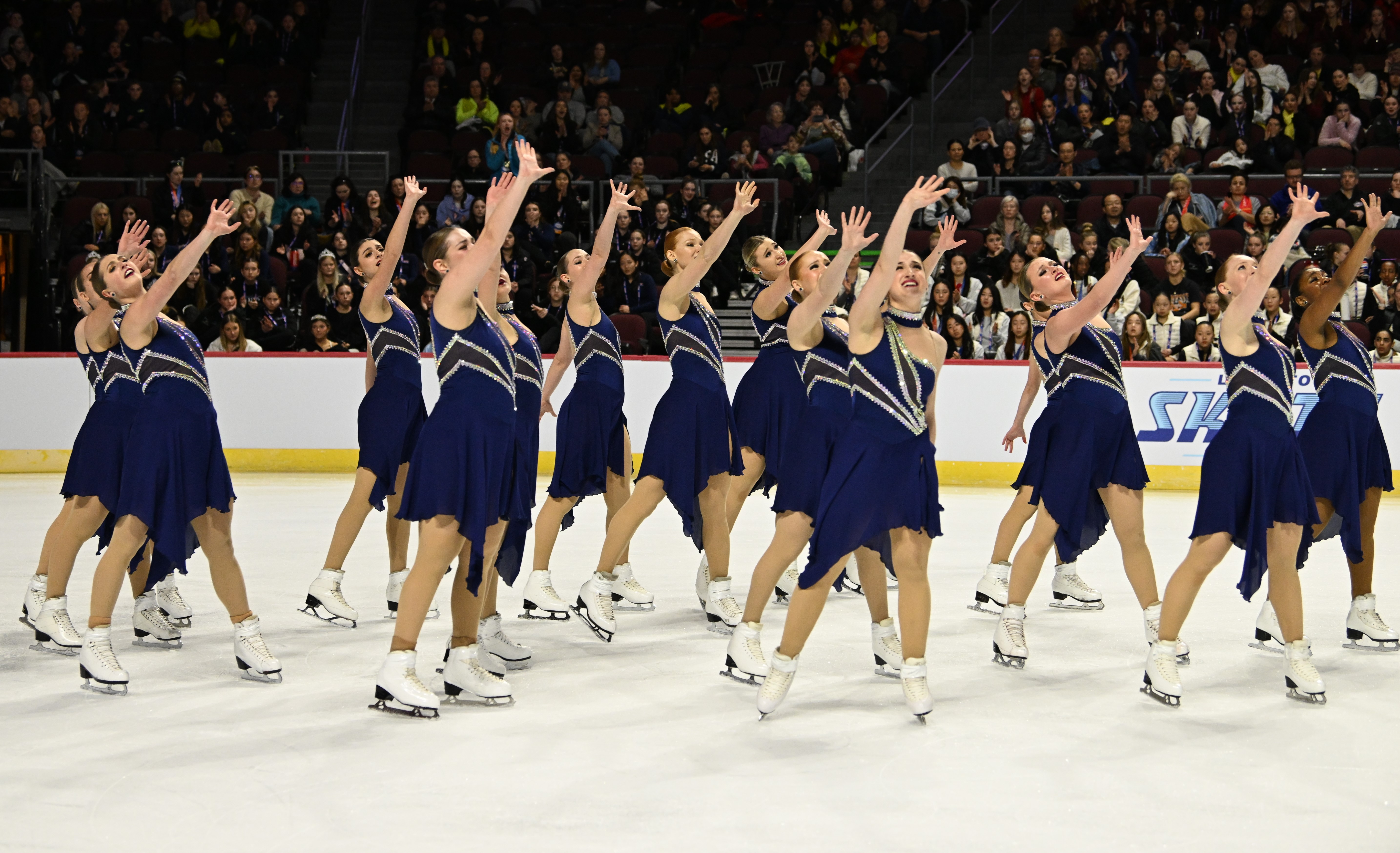  The Skyliners, all standing close together in navy blue dresses with silver lines going diagonally down the dress, reach up across their bodies with their right arm.