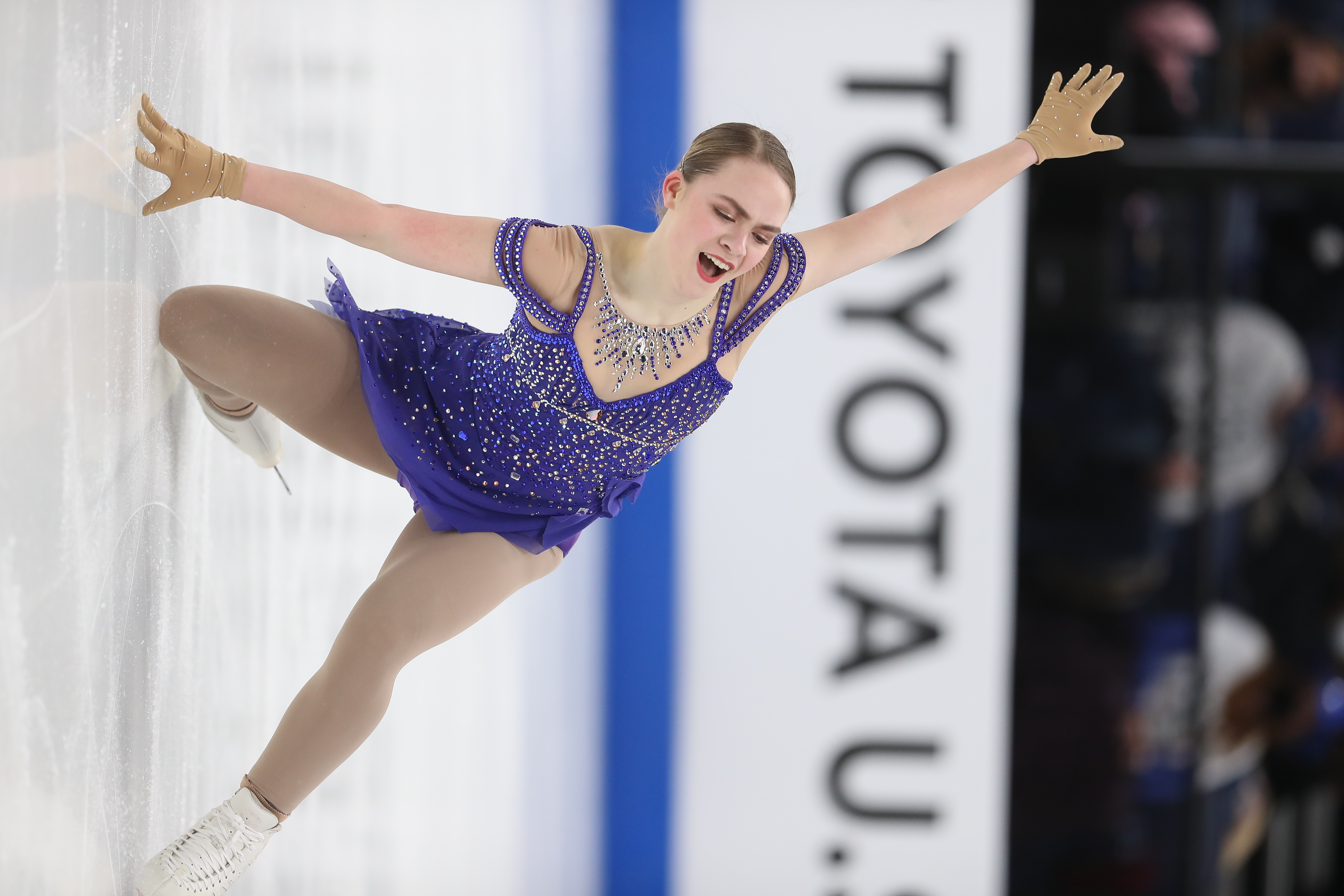 Wren Warne-Jacobson competes at the 2022 U.S. Figure Skating Championships. She is a young white woman with blonde hair tied back in a bun. She is wearing a purple skating costume and kneels on the ice with one hand on the ice and the other raised above her head