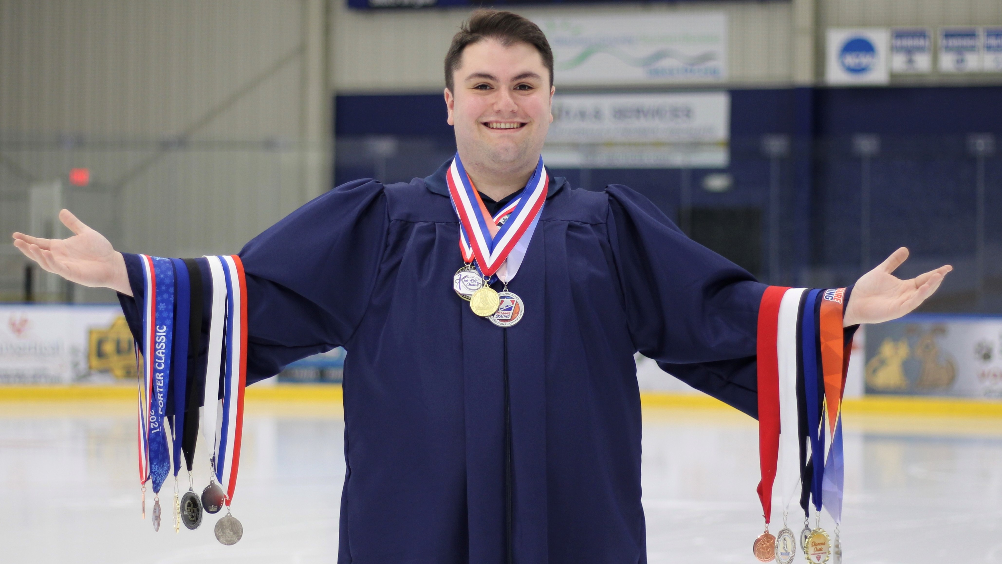 Zak stands in his navy blue graduation gown with his arms outstretched at his side. On each arm hangs several medals from various figure skating competitions. Zak is a young white man with short brown hair