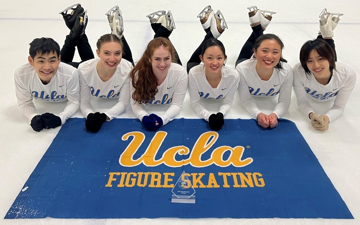 Audrey poses with a few of her teammates on the ice. They all lay on their stomaches with their heads in their hands. A blue UCLA flag is draped on the ground in front of them