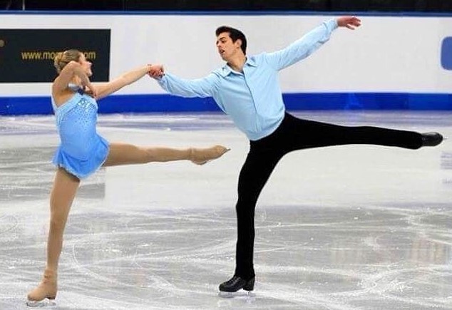 Colin and his partner Kylie skate holding hands with one leg extended behind them. Kylie is wearing a blue skating costume and Colin is wearing a matching long sleeve top and black pants 