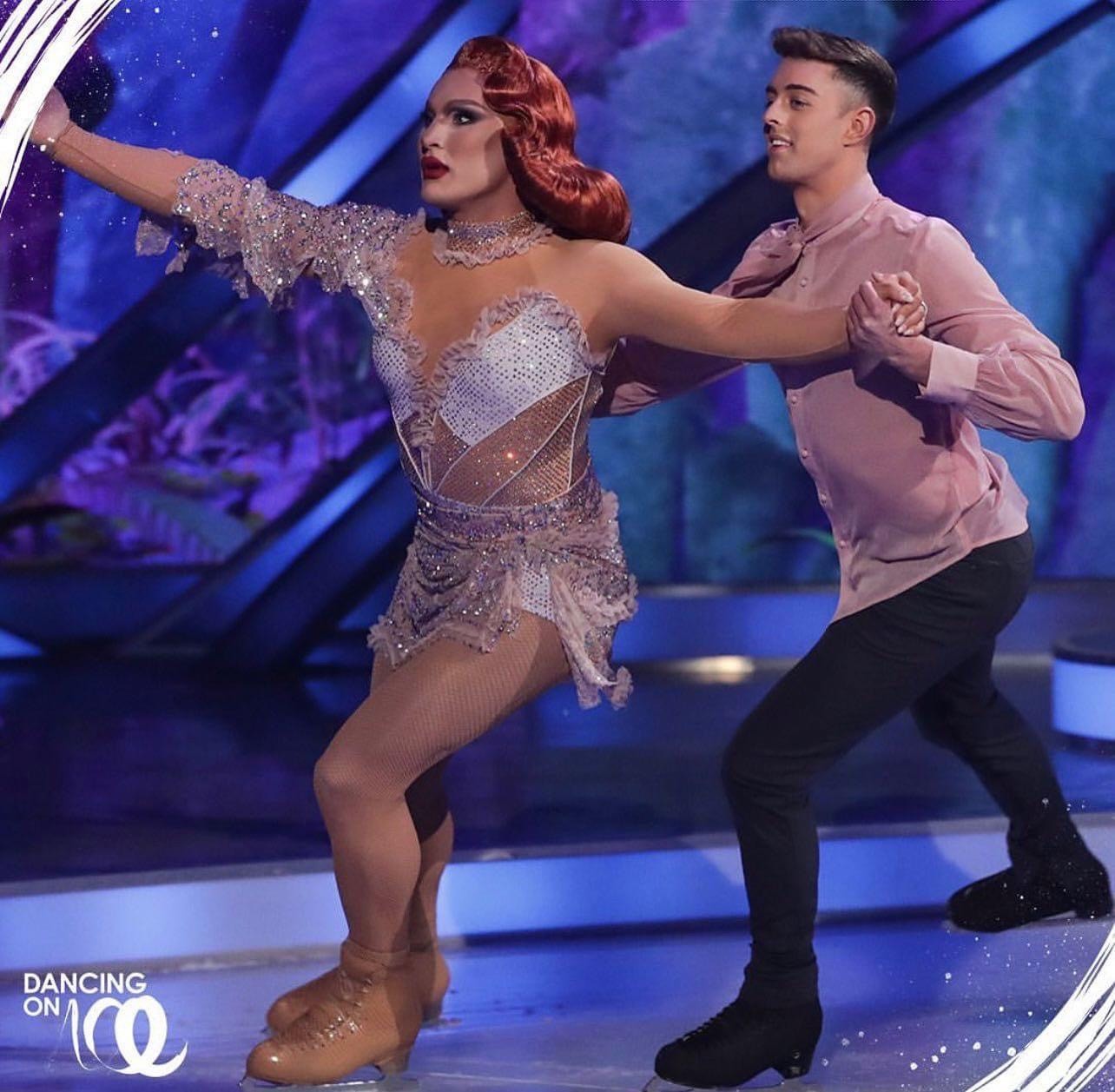 Colin skates while holding the hand of his partner, drag queen The Vivienne. Colin skates wearing a long sleeve pink shirt and The Vivienne has bright red hair and is wearing a nude colored skating costume with sparkles