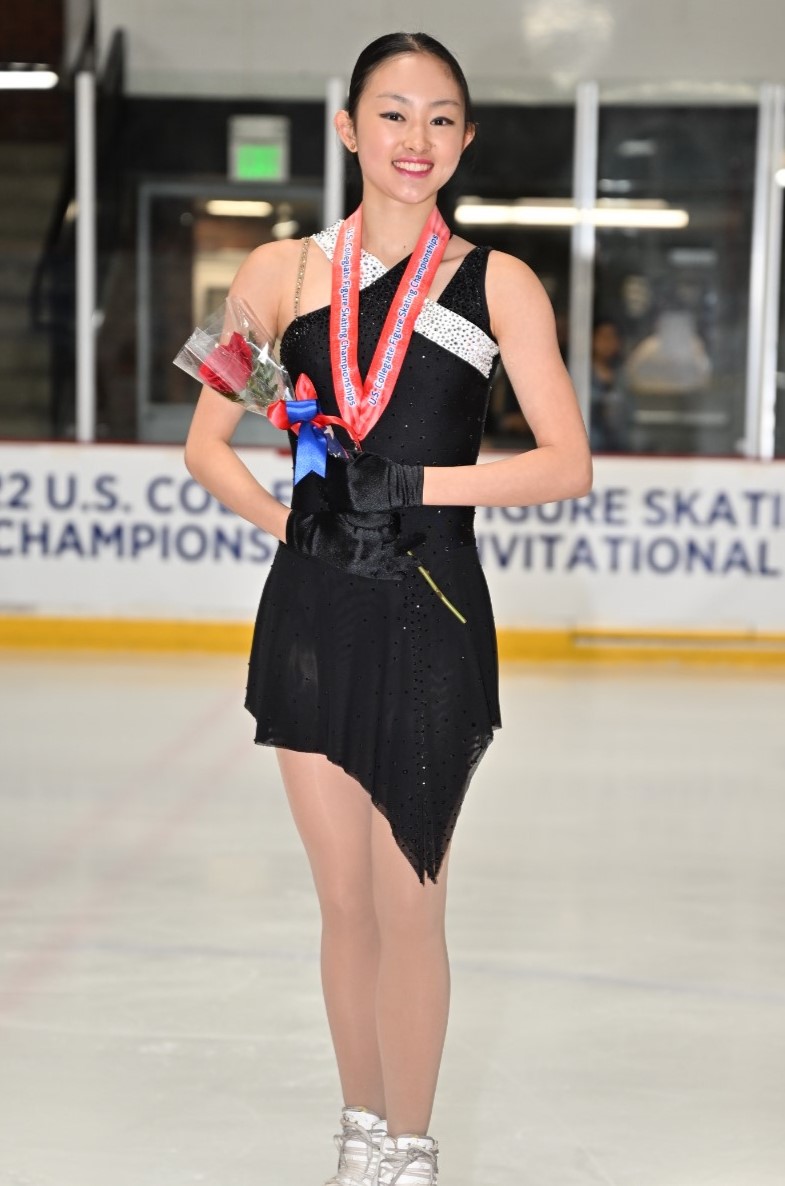 Audrey Lu poses on the podium of the U.S. collegiate championships. She is a young Asian woman wearing a black skating costume with a white stripe across the chest. She is wearing a medal with a red ribbon and holding a single rose with a blue and red ribbon around the stem