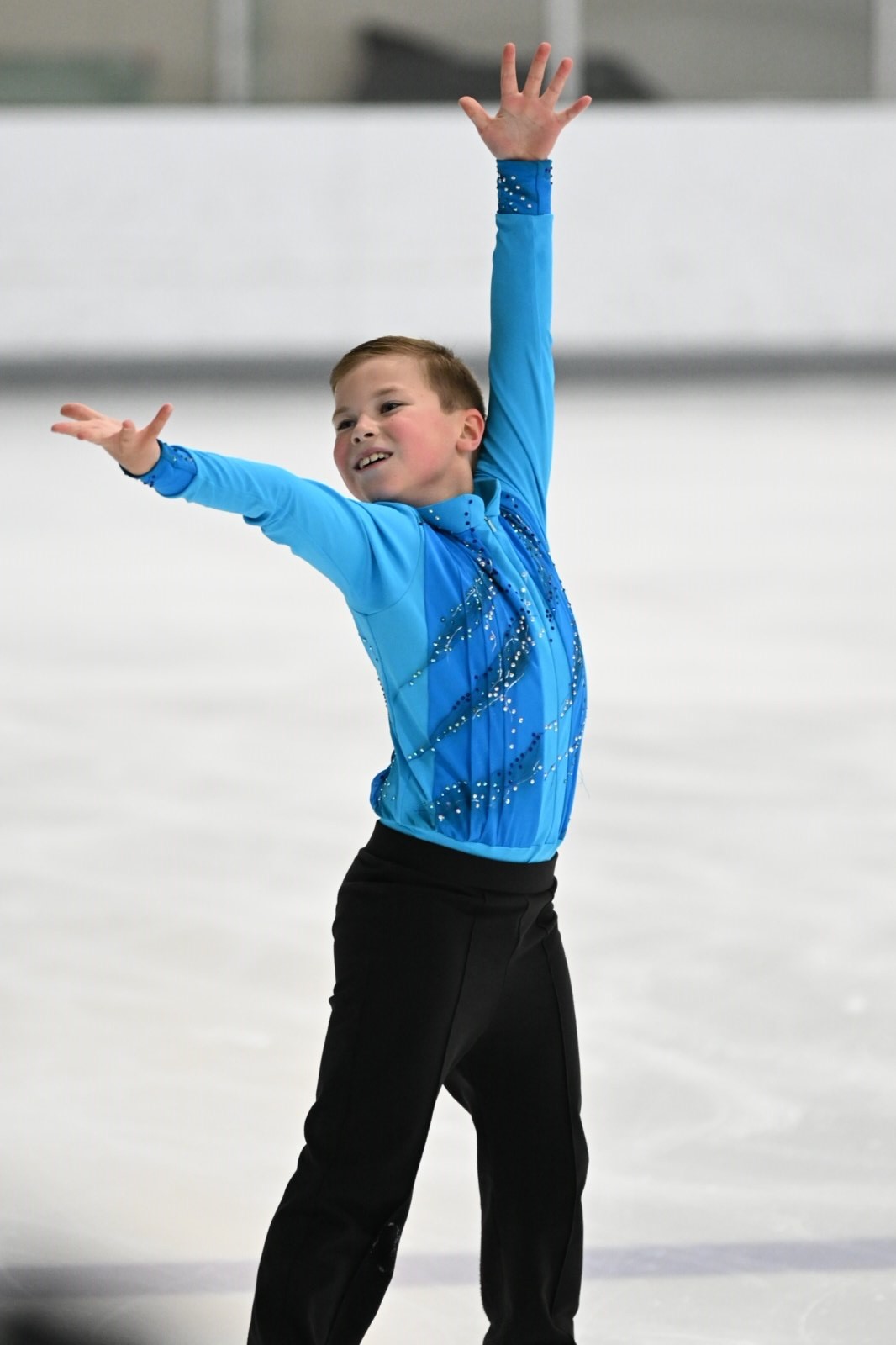 Ryan Lipman competes at the 2023 Excel National Festival. He is a young white boy wearing a long sleeve blue top and black pants. He dramatically strikes a pose with his hands above his head