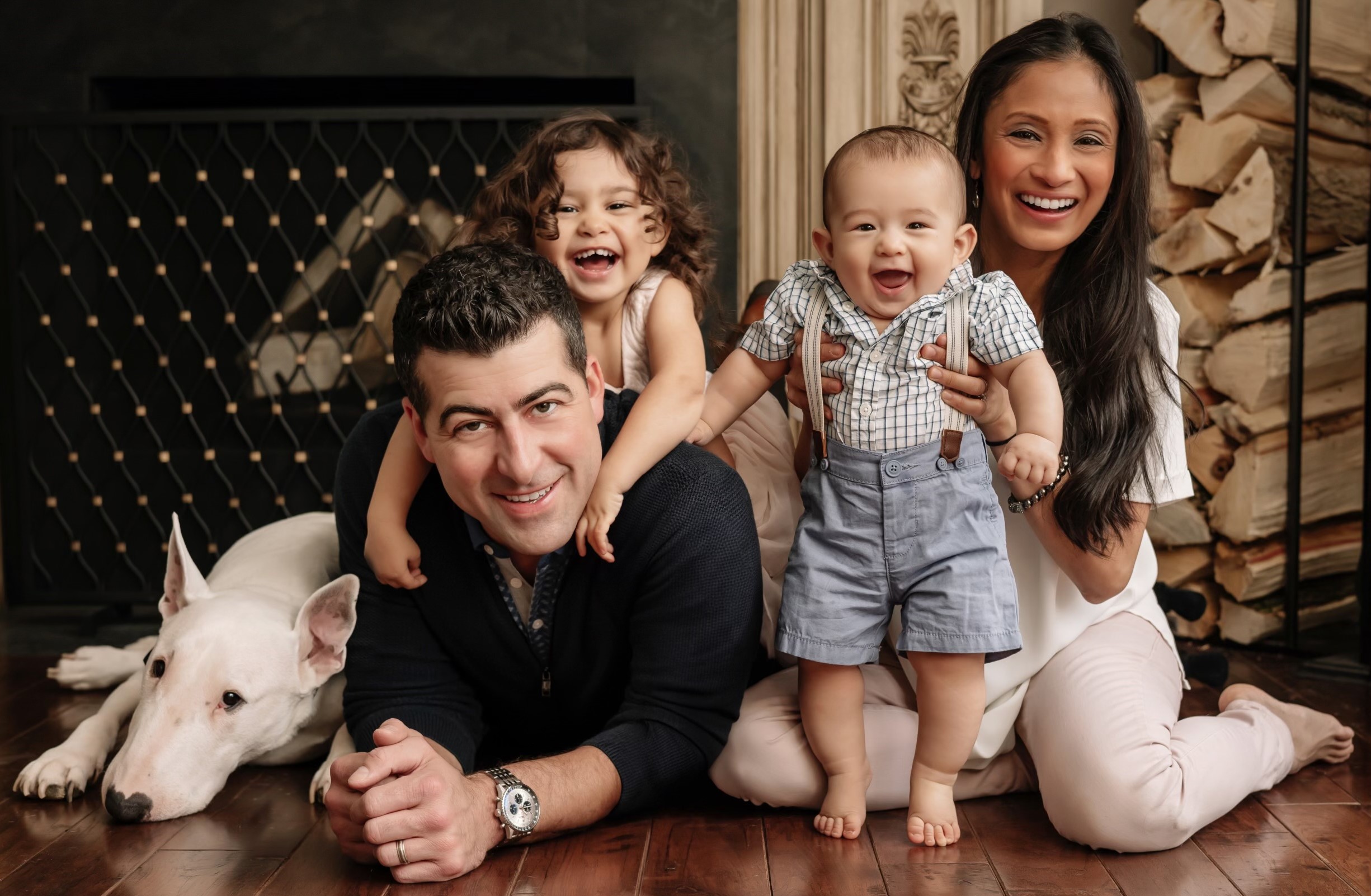 Amanda poses with her family laying on the ground. Her husband lays on his stomach smiling at the camera. Their daughter lays on his shoulders. Amanda sits up supporting her young son as he stands up. The family's white dog lays down to the left of the photo