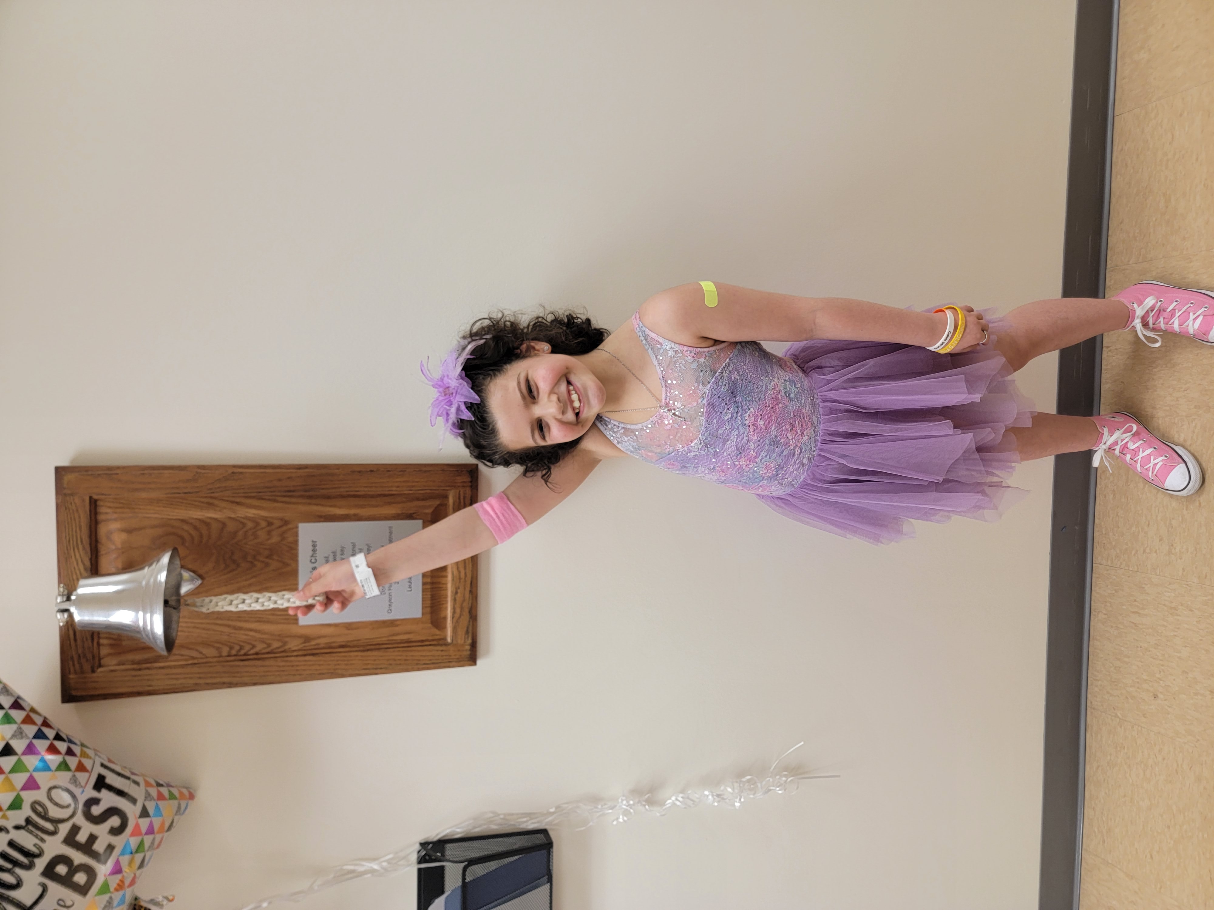 Hannah rings the bell at the hospital signifying that she finished chemotherapy. Hannah is a young girl with short curly brown hair wearing a purple dress and pink converse. She smiles at the camera with her right hand reaching up to hold the rope of a silver bell hanging on the wall. 