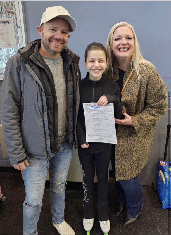 Hannah (center) poses with her coaches holding her pre-preliminary test papers. Hannah is a young girl with short brown hair wearing a black jacket and black pants. To her left is her coach Ben who is an older white man wearing a grey jacket, jeans and a white baseball cap. To her right is her coach Ann who is an older white woman with long blonde hair wearing a leopard print jacket and jeans 