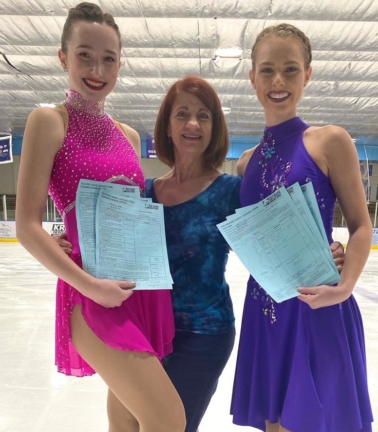 Joelle (left) poses with her coach and another skater after passing her free dance test. Joelle (left) is a young woman with brown hair tied up in a bun wearing a pink skating costume. She stands with an older woman with shoulder length red hair and another skater with blonde hair and she is wearing a purple skating costume