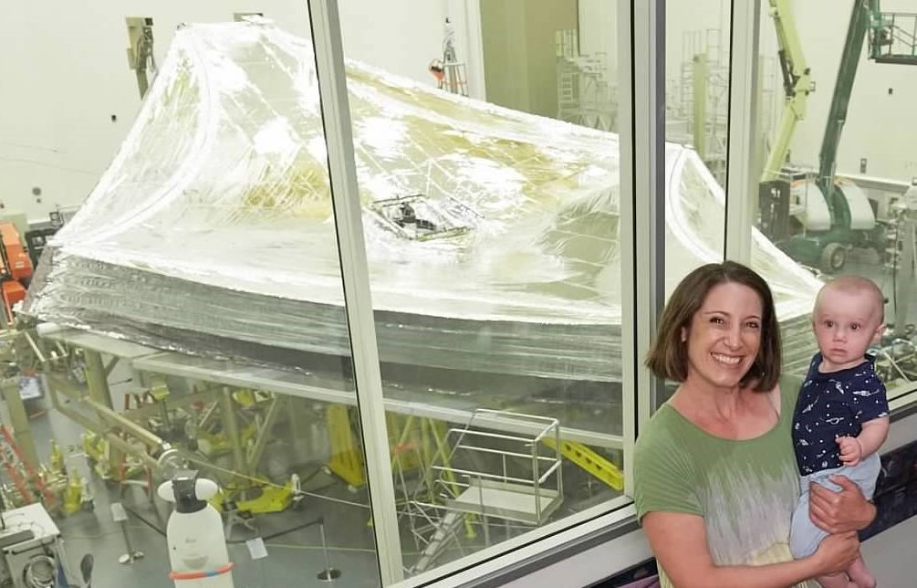 Kelley Ristau stands in front of a large piece of technology for the James Webb Space Telescope while holding a baby