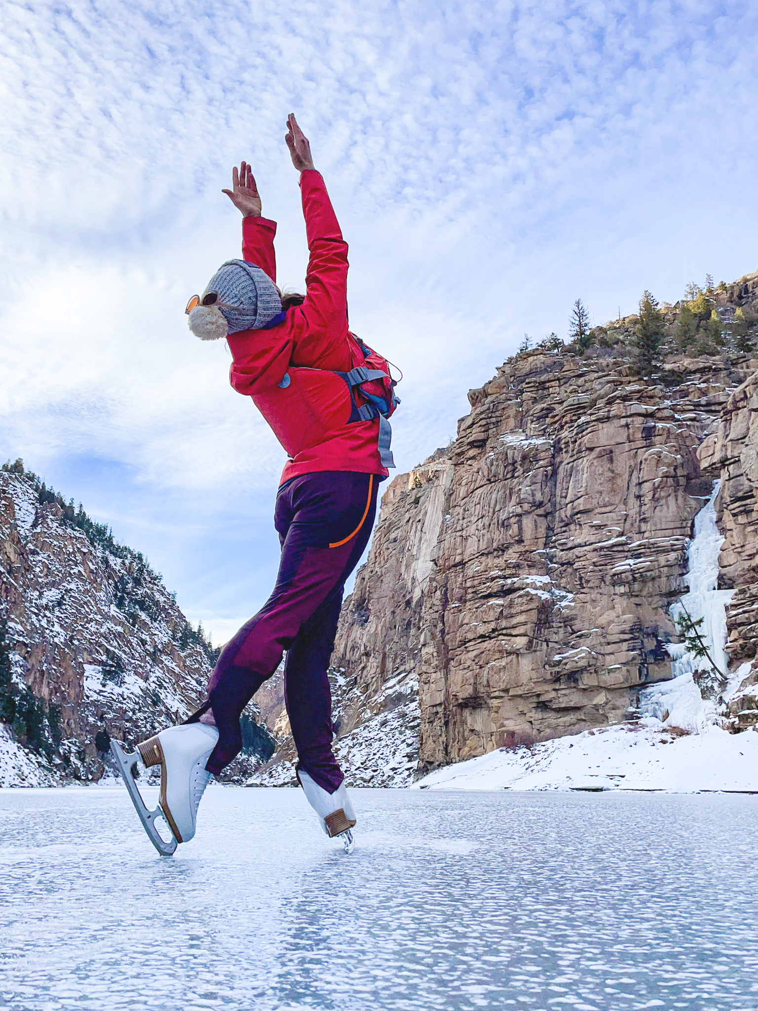 Laura stands on the ice with her arms reaching towards the sky. She is wearing a pink jacket, purple leggings and a blue beanie with her skates