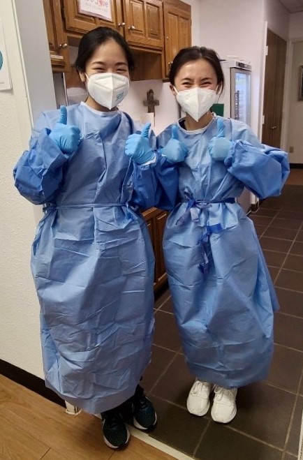 Shin Lei Case poses with a colleague with them both giving a thumbs up to the camera. They are both young Asian women wearing blue surgical gowns and white face masks