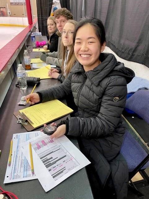 Shin Lei sits at a judges table and smiles at the camera. She is a young Asian woman with black hair tied back in a ponytail. She is wearing a black winter coat