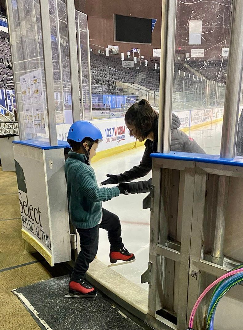 A woman holds the hands of a young skater to help guide him on to the ice. The boy is wearing a blue jacket and blue helmet. The woman has short black hair and is wearing a grey winter coat