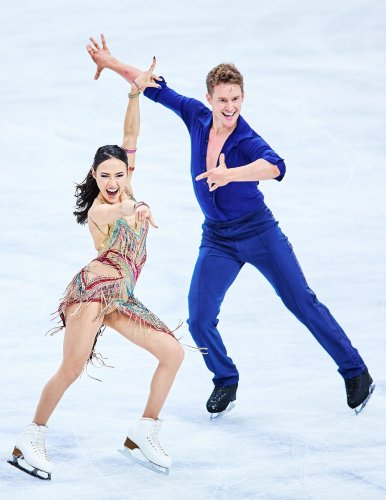 Madison Chock and Evan Bates compete in the rhythm dance at the 2023 Toyota U.S. Figure Skating Championships. Madison (left) is a woman with long black hair wearing a green, pink and purple sparkly skating costume. She has a big smile and her arms are extended outward. Evan stands next to her. He is a white man with short blonde hair wearing a blue top with a deep v neck and matching blue pants. 