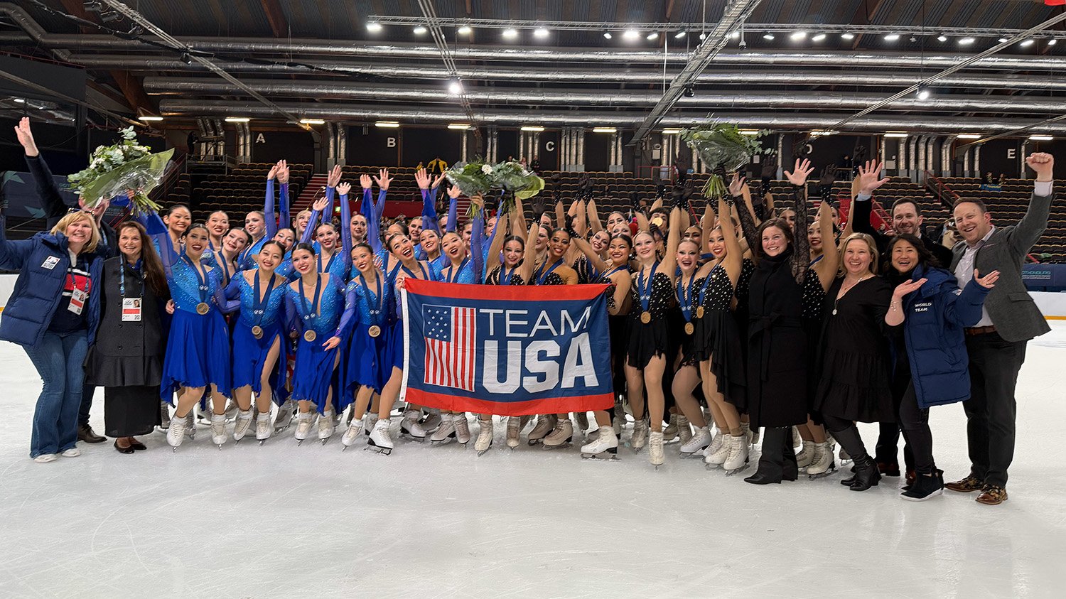 Skyliners junior (left) and Teams Elite (right) celebrate their medals on the ice at the World Junior Championships. The Skyliners wear blue dresses and Teams Elite wears black. Staff stand by them on both sides as they smile and raise their arms up in celebration. They hold a Team USA flag in the middle. 