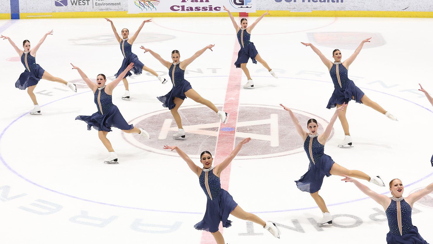 Skyliners skate with their arms outstretched in unison. They all wear matching nay blue skating costumes with a silver collar