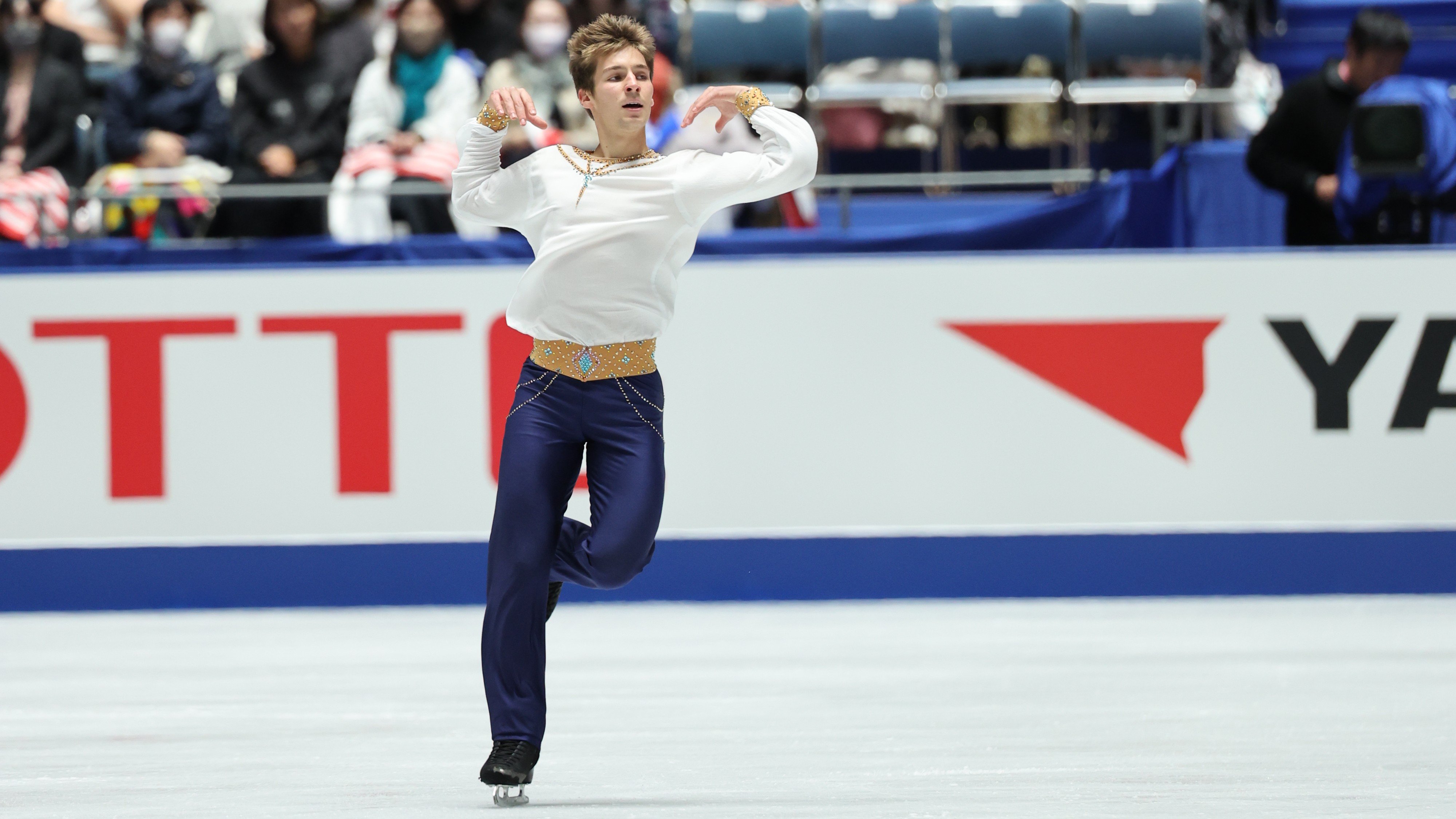 Wearing a white top, tan waistband and blue pants, Andrew Torgashev dazzles the crowd at the NHK Trophy.