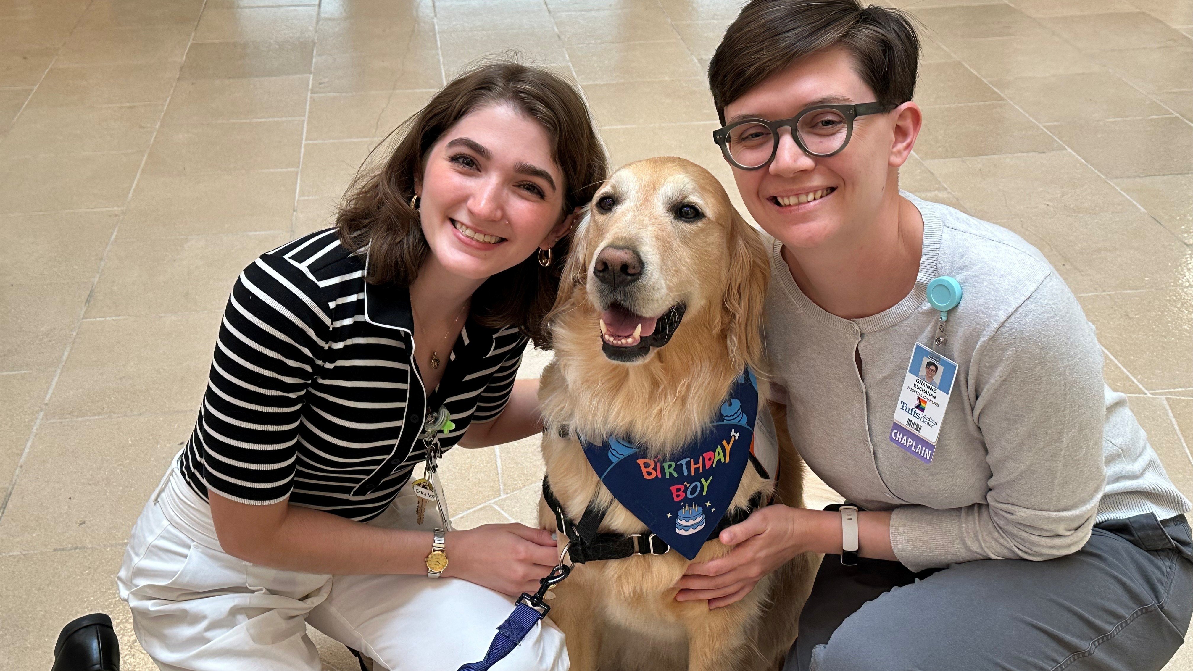 Skyliner Gabriella Romano, a resource specialist at Tuffts Medical Center, is kneeling on the ground with facility dog, Bob, and Gráinne Buchanan, a hospital chaplain. 