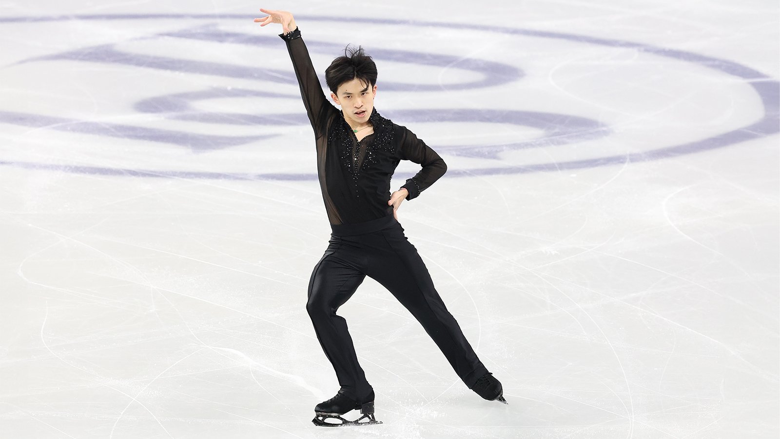 Jimmy Ma strikes a pose on the ice with his left arm extended in the air and his right arm on his hip. He is a young Asian man with short black hair wearing a black long sleeve top with sheer sleeves and jewel detailing down the front and black pants.