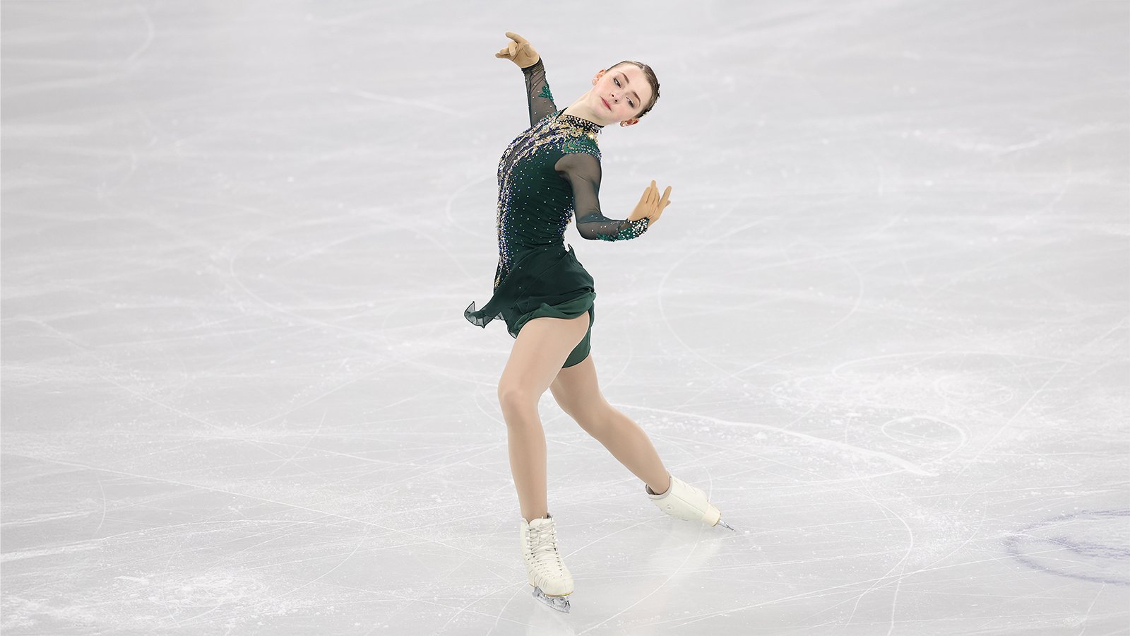 Sarah Everhardt performs her short program wearing a dark green skating costumes with long mesh sleeves. There is jewel detailing down the front of the costume. Sarah is a young woman with long blonde hair tied back in a bun. She stands leaning back with her arms outstretched and her left leg forward. 