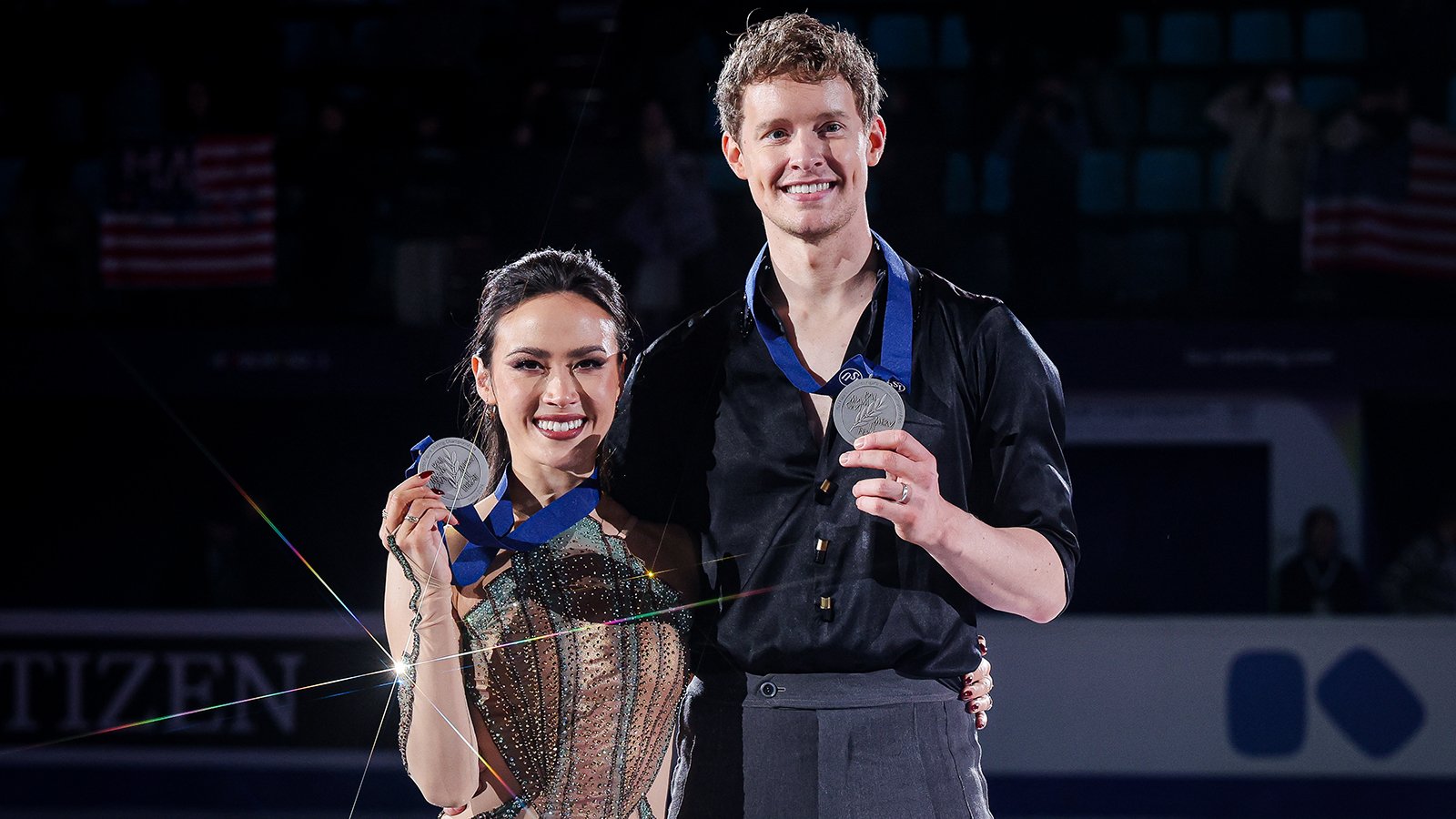 Madison Chock and Evan Bates pose with their Four Continents silver medal. Madison is a young Asian woman with long black hair. She is wearing a copper and teal skating costume designed to resemble the coloring of the Statue of Liberty and the look of the Empire State Building. Evan is a tall young man with short curly blonde hair. He is wearing a black long sleeve button down shirt with the sleeves rolled up to the elbows and dark grey pants. 