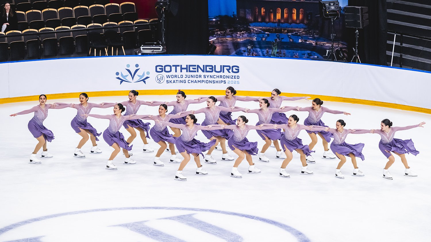 Skyliners junior, wearing purple dresses, skates around the corner of the rink in three lines, each in hold.