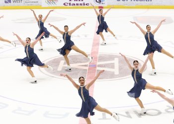 Skyliners skate with their arms outstretched in unison. They all wear matching nay blue skating costumes with a silver collar