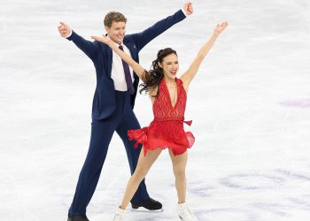 Evan Bates, in a blue tux, white shirt and blue tie, stands behind Madison Chock, who is wearing a red V-neck dress, on the ice. They both have their hands in the air in a Y shape and smile.