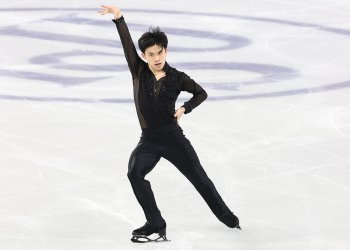 Jimmy Ma strikes a pose on the ice with his left arm extended in the air and his right arm on his hip. He is a young Asian man with short black hair wearing a black long sleeve top with sheer sleeves and jewel detailing down the front and black pants.