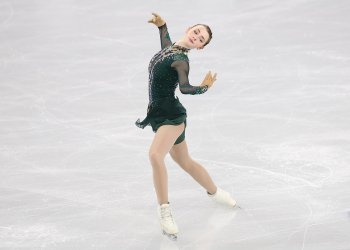 Sarah Everhardt performs her short program wearing a dark green skating costumes with long mesh sleeves. There is jewel detailing down the front of the costume. Sarah is a young woman with long blonde hair tied back in a bun. She stands leaning back with her arms outstretched and her left leg forward. 