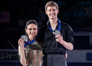 Madison Chock and Evan Bates pose with their Four Continents silver medal. Madison is a young Asian woman with long black hair. She is wearing a copper and teal skating costume designed to resemble the coloring of the Statue of Liberty and the look of the Empire State Building. Evan is a tall young man with short curly blonde hair. He is wearing a black long sleeve button down shirt with the sleeves rolled up to the elbows and dark grey pants. 