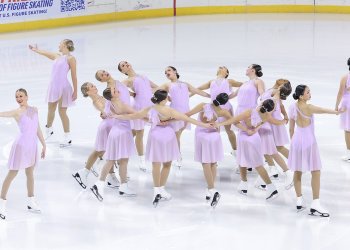 The Hockettes stand in a circle looking outward with their hands around each other's waists. Four skaters stand outside of the circle looking outward with one arm outstretched. They are all wearing matching light pink one-shoulder skating costumes