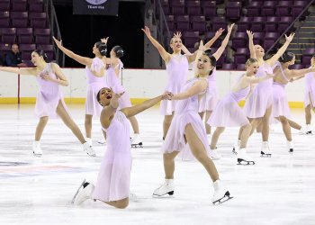 The Hockettes perform wearing coordinating light purple skating costumes. All athletes are standing except one who is on their knees. They all have smiles on their face and their arms raised in the air