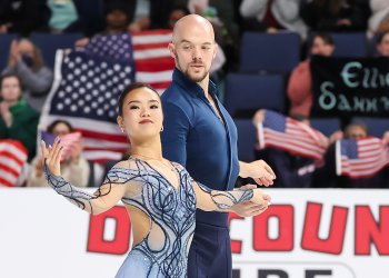 Ellie Kam and Danny O'Shea, side by side, holding hands, compete at 2024 Skate America, with American flags in the background