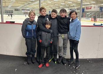 The six members of the Boy Crew stand rinkside. They are six young boys wearing winter jackets. Five stand next to each other with their arms around each other while one stands in front. 