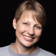 Headshot of a middle aged Caucasian woman smiling. She has short blonde hair and is wearing a grey shirt