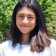 Headshot of an Indian woman smiling. She has shoulder length black hair and is wearing a white long sleeve shirt. Trees are behind her in the background