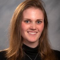 Headshot of a Caucasian woman smiling. She has light brown hair and she is wearing a black turtleneck with a dainty necklace. 