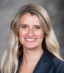 Photo of a Caucasian woman smiling. She has long blonde hair and is wearing a navy blue blazer.