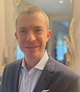 Headshot of Tim Goebel. Tim is a man with a blonde buzz cut. He is wearing a grey suit jacket with a white shirt on underneath