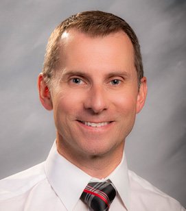 Headshot of a Caucasian man smiling. He has short blonde hair and is wearing a white collared shirt and plaid tie. 