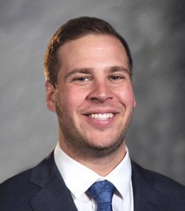 Headshot of a Caucasian man smiling. He has short blonde hair and is wearing a white collared dress shirt, navy blue suit jacket and blue tie