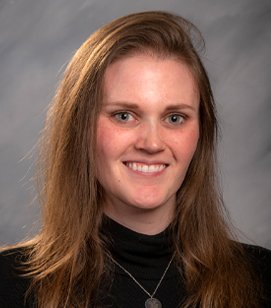 Headshot of a Caucasian woman smiling. She has light brown hair and she is wearing a black turtleneck with a dainty necklace. 