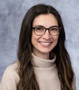 Headshot of Sarah Tyler. She is a Caucasian woman with long brown hair. She is wearing a tan colored turtleneck and glasses