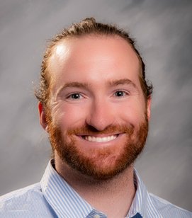 Headshot of a Caucasian man smiling. He has red hair and a beard. He is wearing a light blue collared shirt. 