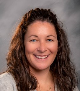 Headshot of a Caucasian woman smiling. She has long brown hair and is wearing a grey shirt.